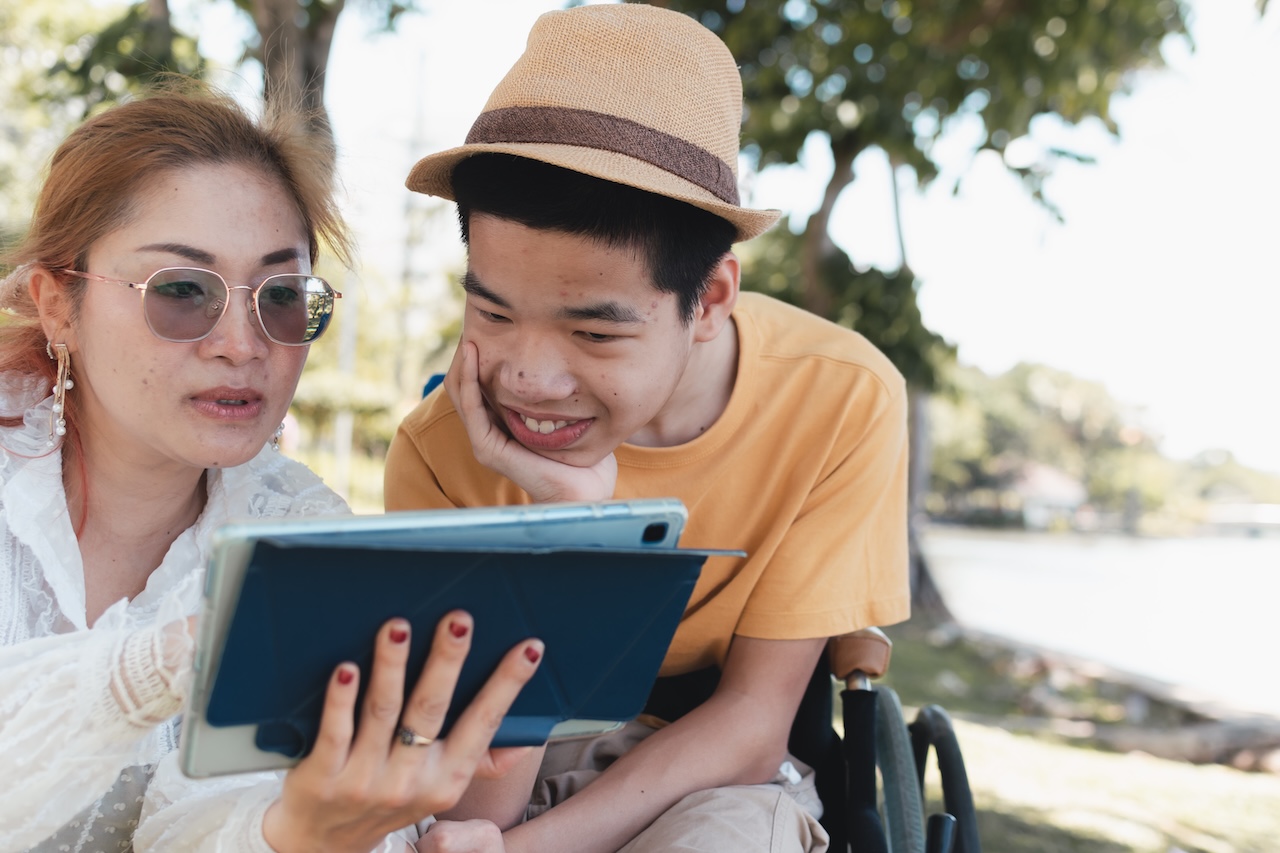 a woman and her son are sat outside. the son is in a wheelchair, and is wearing a fedora-style hat. they are both looking down at a digital tablet screen. the mother is pointing at something and the son is smiling at the screen. the mother is wearing sunglasses.