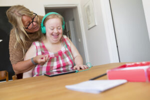 a young woman is sat at a table indoors looking at a digital tablet. she has headphones on and is smiling. her mother is behind her with an arm around her pointing to the tablet.