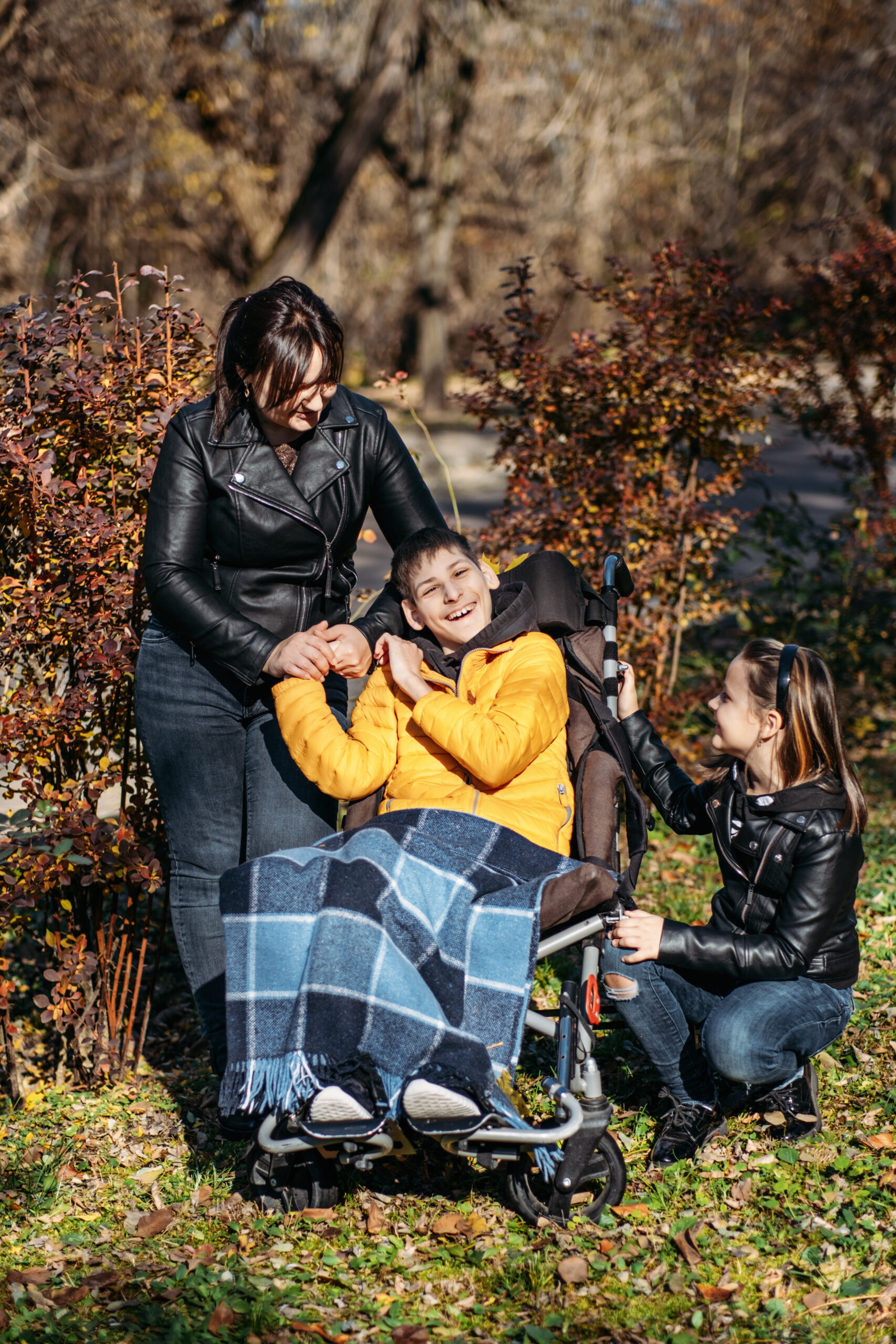 a young man is sat outdoors in his wheelchair with a blanket over his legs. his mother is stood behind him with an arm over his shoulders. his sister is knelt at his other side with a hand on his wheelchair. they are all smiling.