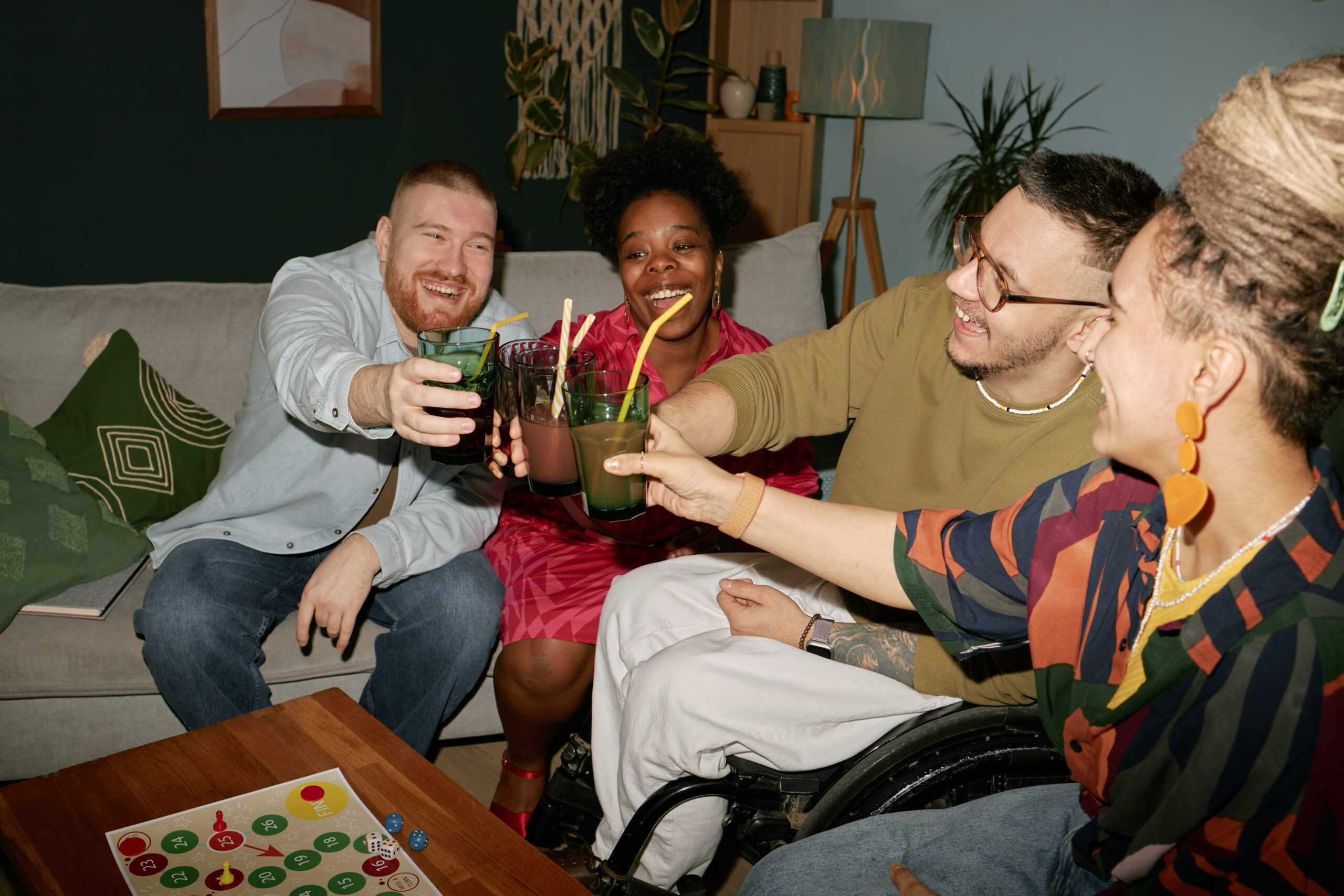 a group of friends are sat on sofas celebrating, and cheers-ing their drinks. the man on the middle right side is in a wheelchair. there is a board game on the table. they are all smiling.
