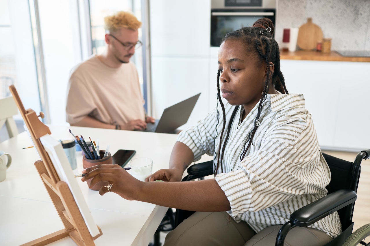 a woman in a wheelchair is sat at a desk. a man on a laptop is in the background. in front of the woman is an easel with a canvas on it. next to her there is a glass of water and a pot of brushes. she is painting on the canvas. she looks concentrated.