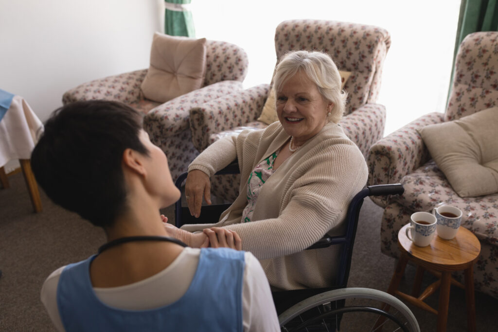an elderly woman is sat indoors in a wheelchair. behind her, there are three patterned armchairs. a healthcare worker is talking to her, and they are both smiling. on the table to the right, there are two mugs.