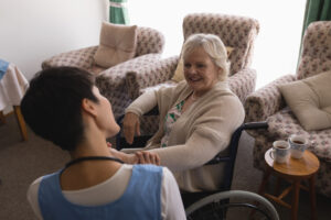 an elderly woman is sat indoors in a wheelchair. behind her, there are three patterned armchairs. a healthcare worker is talking to her, and they are both smiling. on the table to the right, there are two mugs.