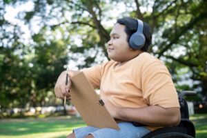 a young man in a wheelchair is sat outside. he has headphones on. in his hands there is a clipboard and a pencil. he is looking forwards and smiling.