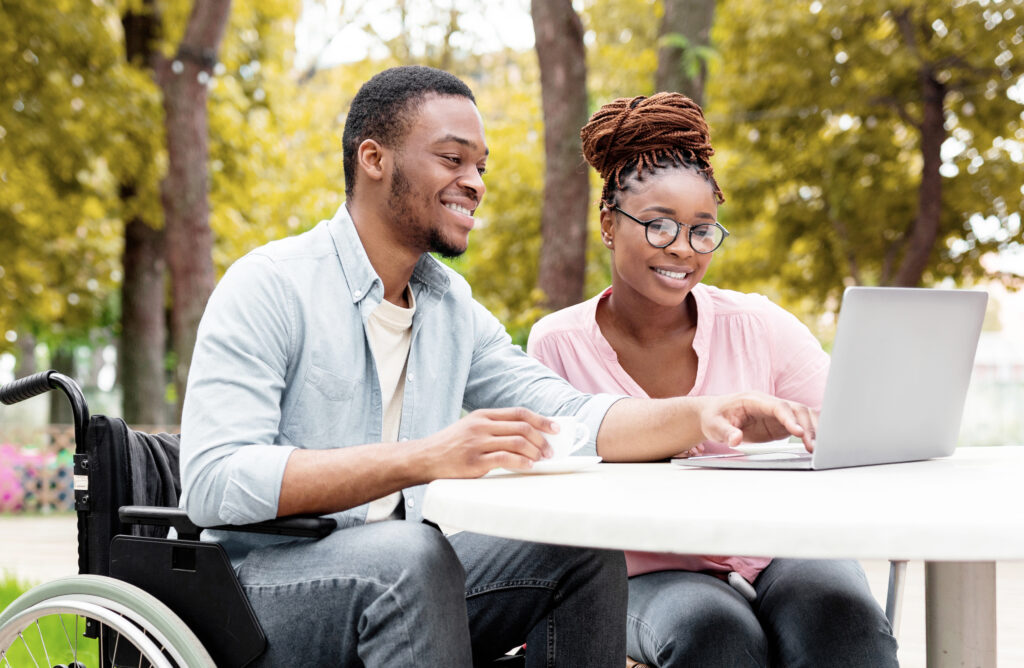 a woman and a man are sat outdoors at a table. there are trees in the background. the man is in a wheelchair. in front of them, there is an open laptop which the man is typing on. he is also holding a mug in his left hand. they are both looking at the laptop screen and smiling.