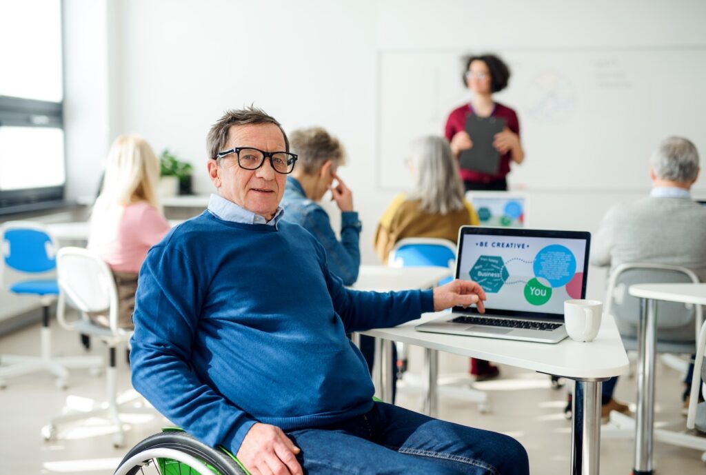 An elderly man with a wheelchair is in focus while there is a classroom meeting in the background out of focus. He has a laptop open to a slide saying "Be Creative".