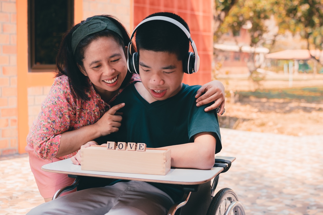 a young man and his mother are sat outside playing with blocks. the young man is in a wheelchair with a desk in front of him, and has headphones on. the wooden blocks are laid out horizontally on a wooden box. they spell out love. the mother is looking down at the blocks and pointing at the boy. they are both smiling.