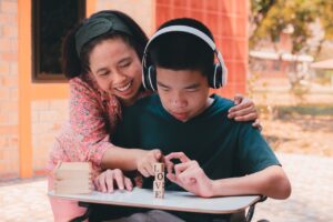 a young man and his mother are outside. he is sat at a desk playing with blocks that vertically spell out love. he is wearing headphones and smiling. his mother has her arm wrapped around him and one hand pointing at the blocks. she is also smiling.