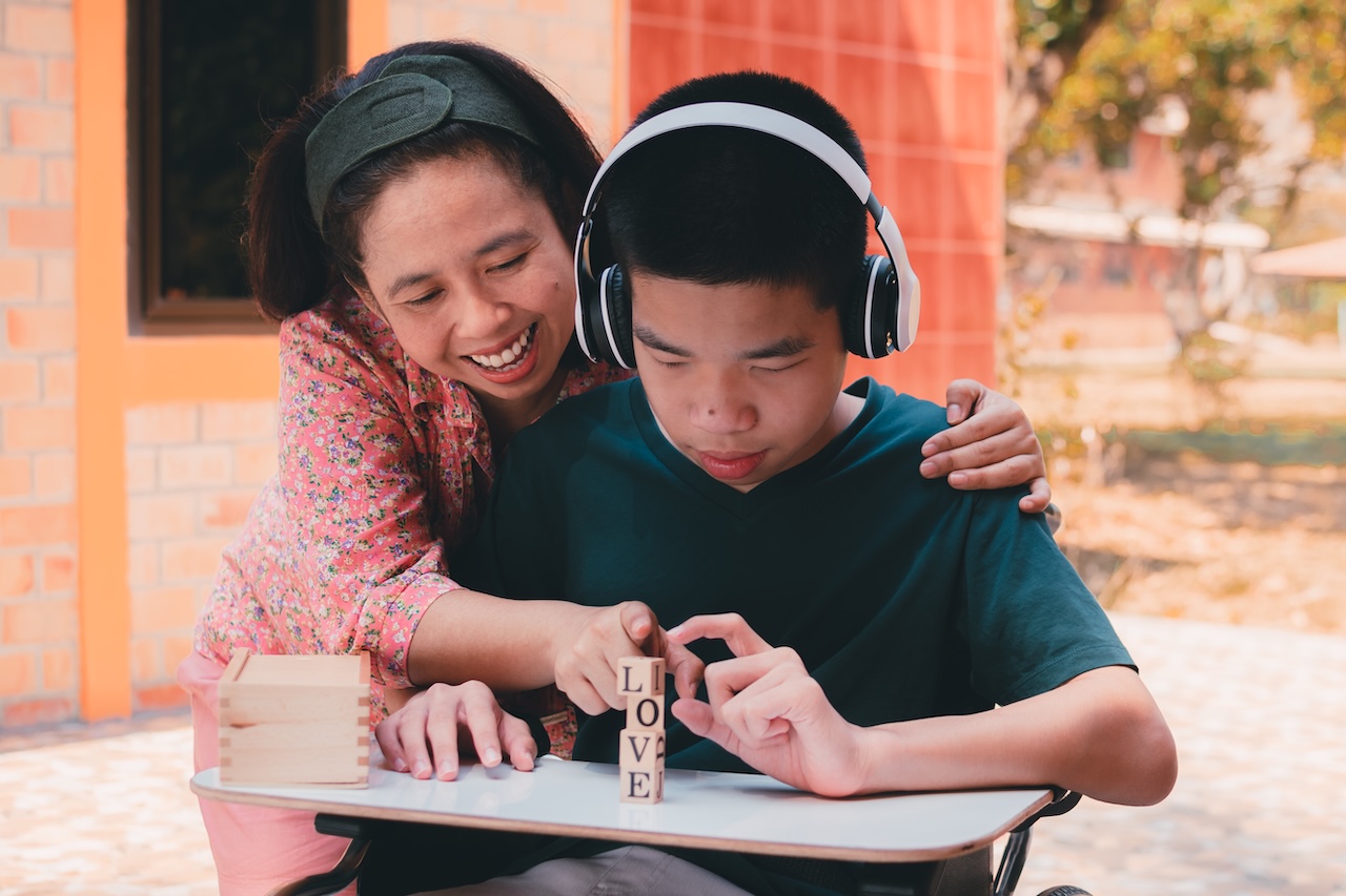 a young man and his mother are outside. he is sat at a desk playing with blocks that vertically spell out love. he is wearing headphones and smiling. his mother has her arm wrapped around him and one hand pointing at the blocks. she is also smiling.
