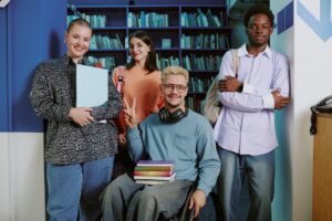 a group of 4 people are stood together in a library. they are all posing. on the left, there are two women, the one on the far left is holding a notebook. on the right there are two men. the man on the far right has his arms crossed. the man in the middle is in a wheelchair and has a stack of books in his lap.