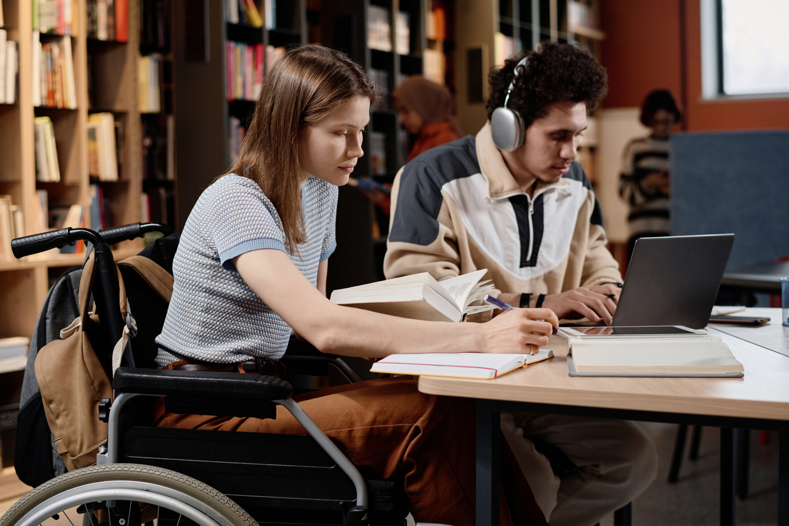 a young woman and man are sat at a desk in a library. the young woman is in a wheelchair. she is looking through a book and writing in a notebook. the man next to her has headphones on and is typing on a laptop.