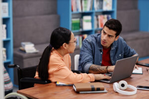 a young woman and man are sat at a desk in a library. the young woman is in a wheelchair. there is a laptop and a notebook on the desk. they are both engaging in a discussion.