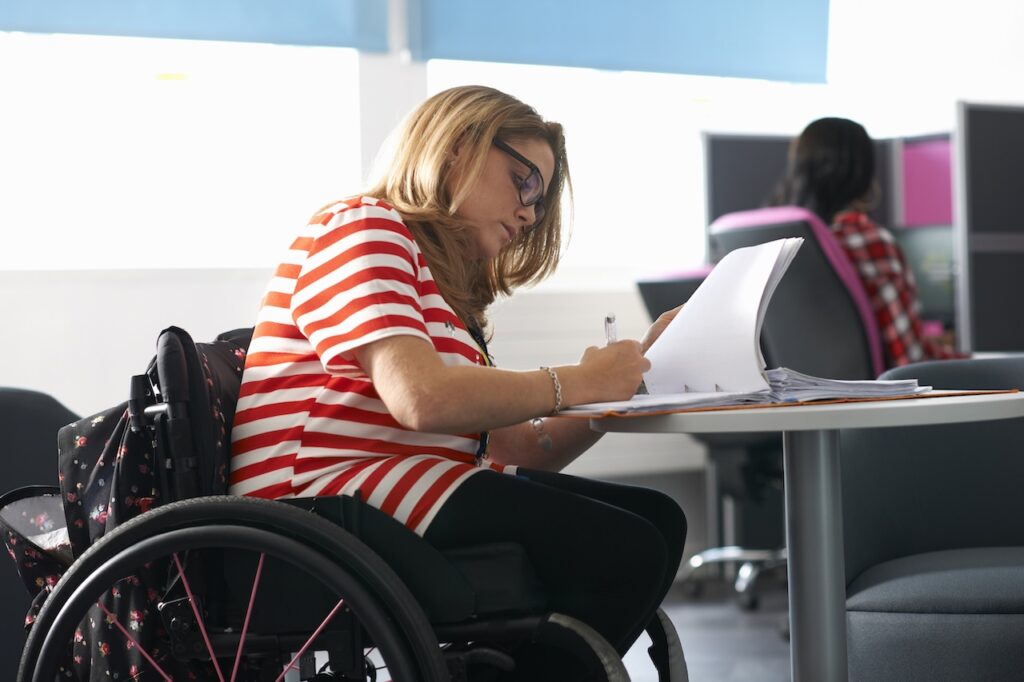 A young woman is in a wheelchair and is concentrating on writing in her notebook which is on a table. She has glasses. There is someone on a computer in the background.