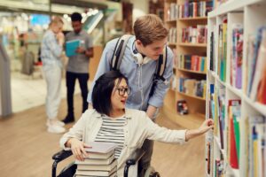 a young man and woman are in a library picking out books. the woman is in a wheelchair, and has a stack of books on her lap. she is selecting a book from the shelf. the young man is pushing her, and has a backpack and headphones on. they are both smiling.