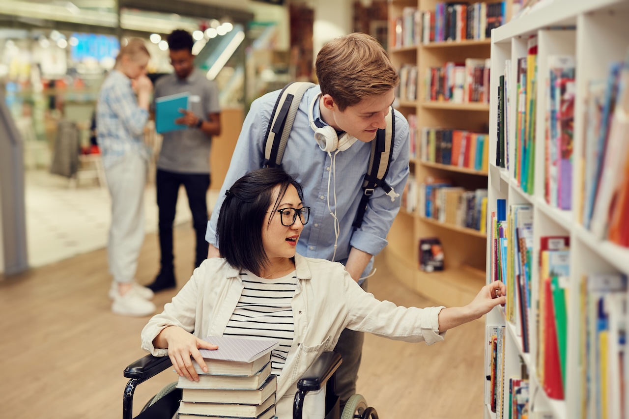 a young man and woman are in a library picking out books. the woman is in a wheelchair, and has a stack of books on her lap. she is selecting a book from the shelf. the young man is pushing her, and has a backpack and headphones on. they are both smiling.