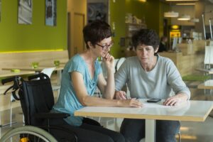 two women are sat a table in a cafe. they are both looking down at a piece of paper. the woman on the right has a pair of glasses and a phone in front of her. the woman on the left is in a wheelchair.