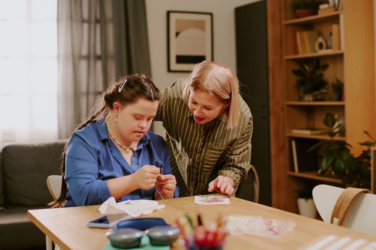 a young woman with down-syndrome is sat at a table threading a needle. her mother is stood beside her advising her what to do. they appear to be doing some kind of embroidery. the mother is smiling.