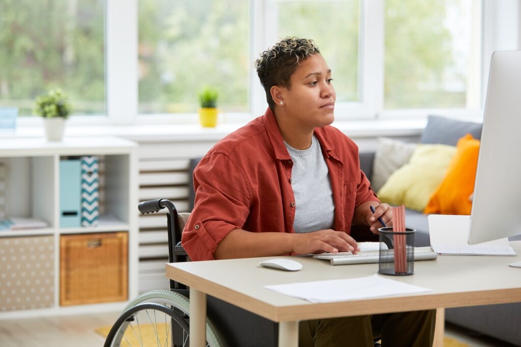 A young person in a wheelchair is sat at a desk. They are typing on a keyboard and holding a pen in one hand. There is a notebook to the side. In front of them, there is a large computer screen.