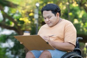 a young man in a wheelchair is sat outdoors on a sunny day. he is holding a coffee cup and a clipboard in one hand, and a pen in the other. he is smiling and concentrating on the clipboard.