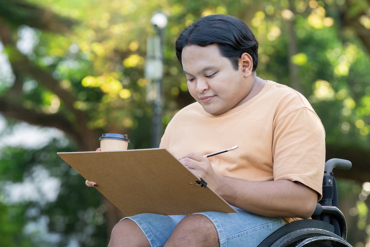 a young man in a wheelchair is sat outdoors on a sunny day. he is holding a coffee cup and a clipboard in one hand, and a pen in the other. he is smiling and concentrating on the clipboard.