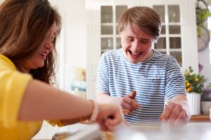 a woman, and a man with down-syndrome are stood in the kitchen baking. they are putting mixture into a muffin tin. both are smiling.