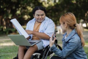 a young man in a wheelchair is sitting outside. he has a laptop on his lap and is holding a notebook. a young woman is crouched next to him. he is pointing to the notebook and looking at her.