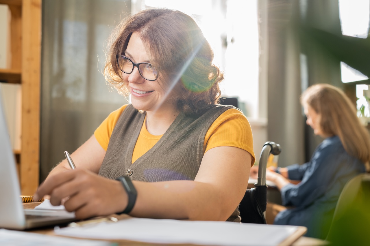 a woman in a wheelchair is sitting at a library desk on a sunny day. she is holding a pen in her hand and is smiling down at a laptop screen.