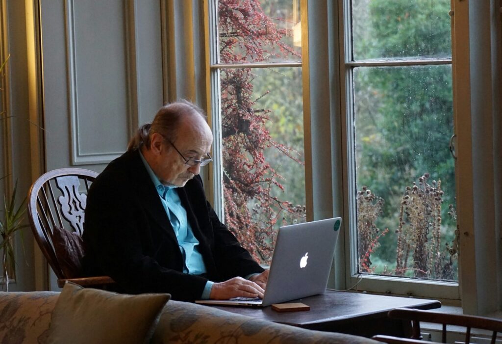 An elderly man with glasses is sat by a window on his own and is staring at an open laptop on a table.