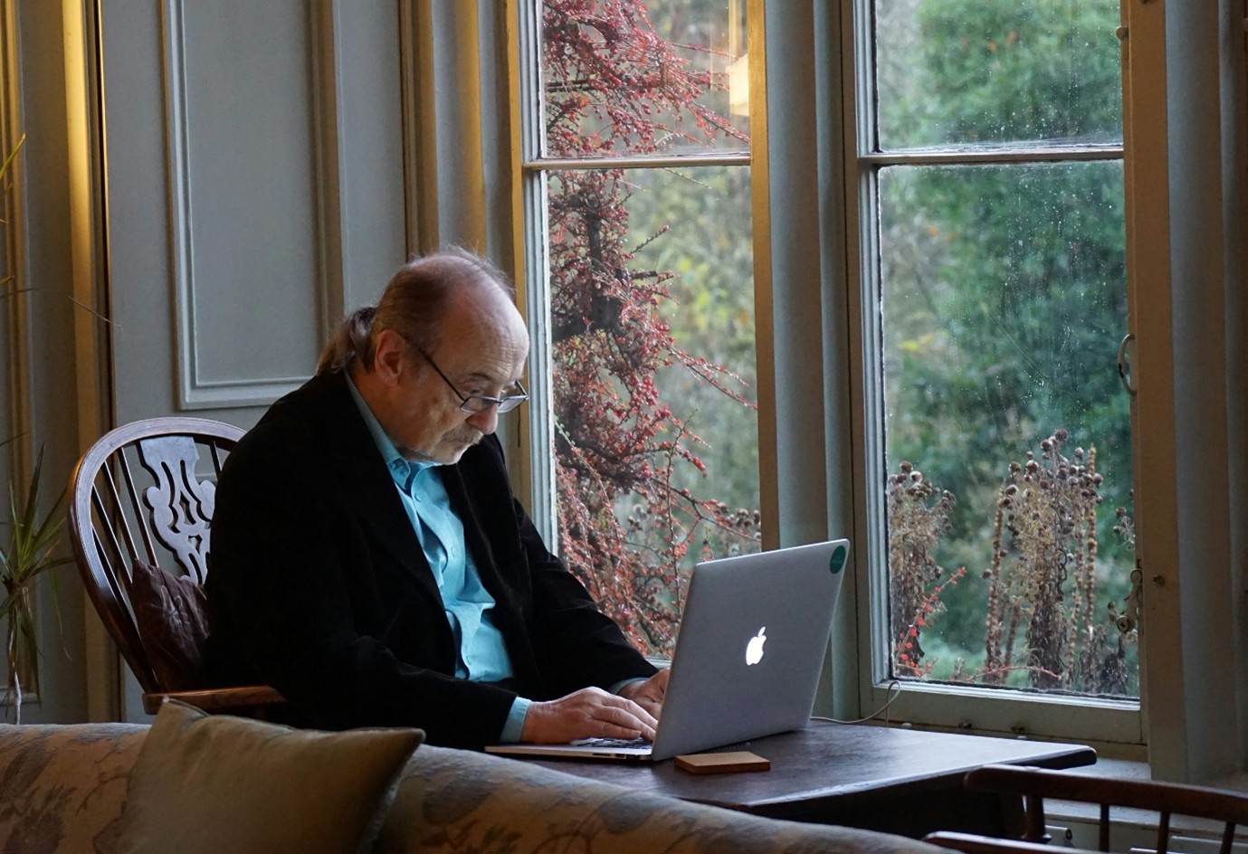 an elderly man is sat at a table by a window, with a laptop in front of him. he is typing, and focused on the laptop screen.