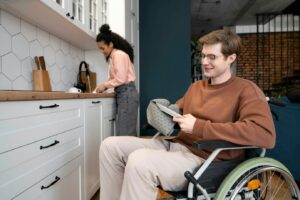 a young couple are washing dishes in the kitchen. a young woman is at the sink, and a man in a wheelchair is drying a plate with a tea towel. they are both smiling.