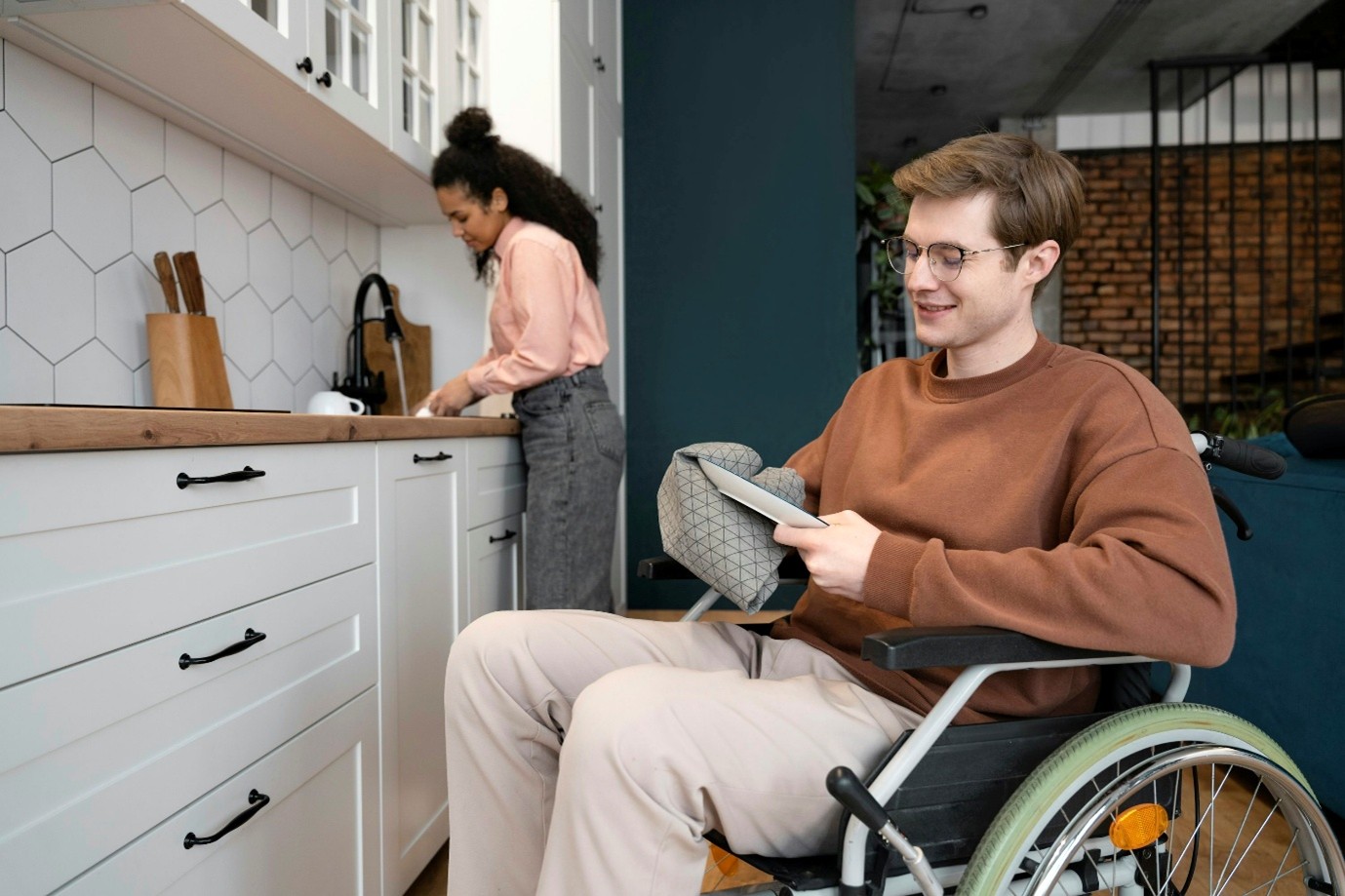 a young couple are washing dishes in the kitchen. a young woman is at the sink, and a man in a wheelchair is drying a plate with a tea towel. they are both smiling.