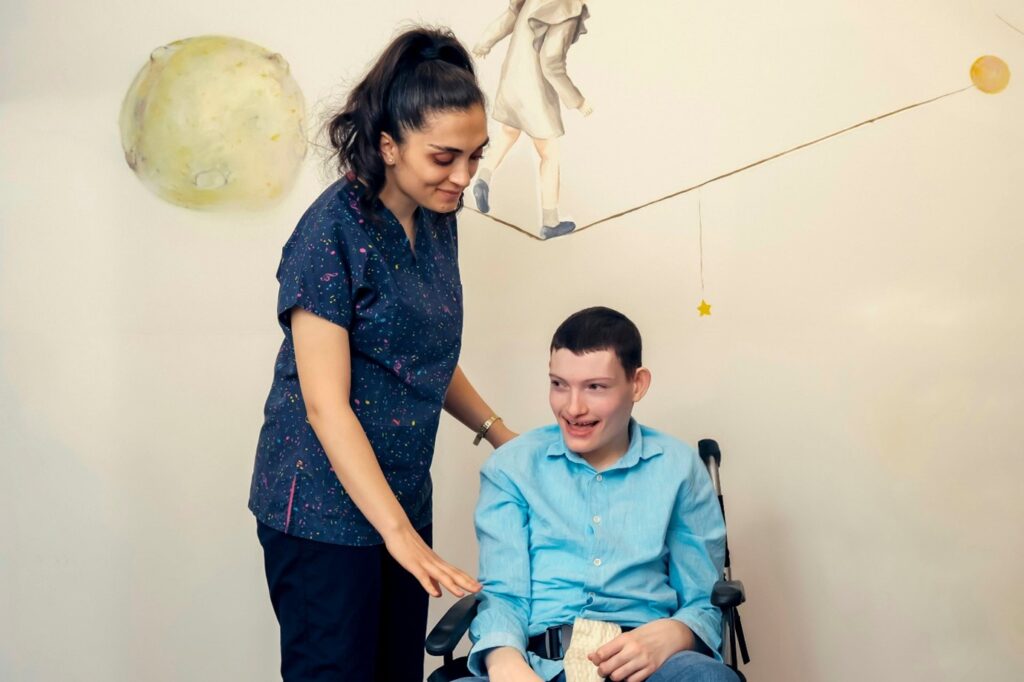 a young man in a wheelchair is with his carer, who has her arm around him while standing up. they are both smiling.