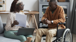 a man and a woman are sitting inside having a discussion. the woman is holding a piece of paper and has a laptop on her lap. the man is a wheelchair-user.