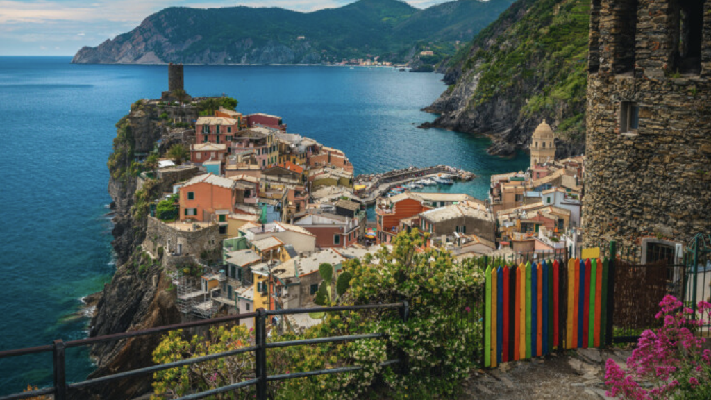 a view from a high point of a colourful european city by the coast. there is a rainbow fence surrounded by thinner black fences in the foreground. in the background there are green hills and cliffs.