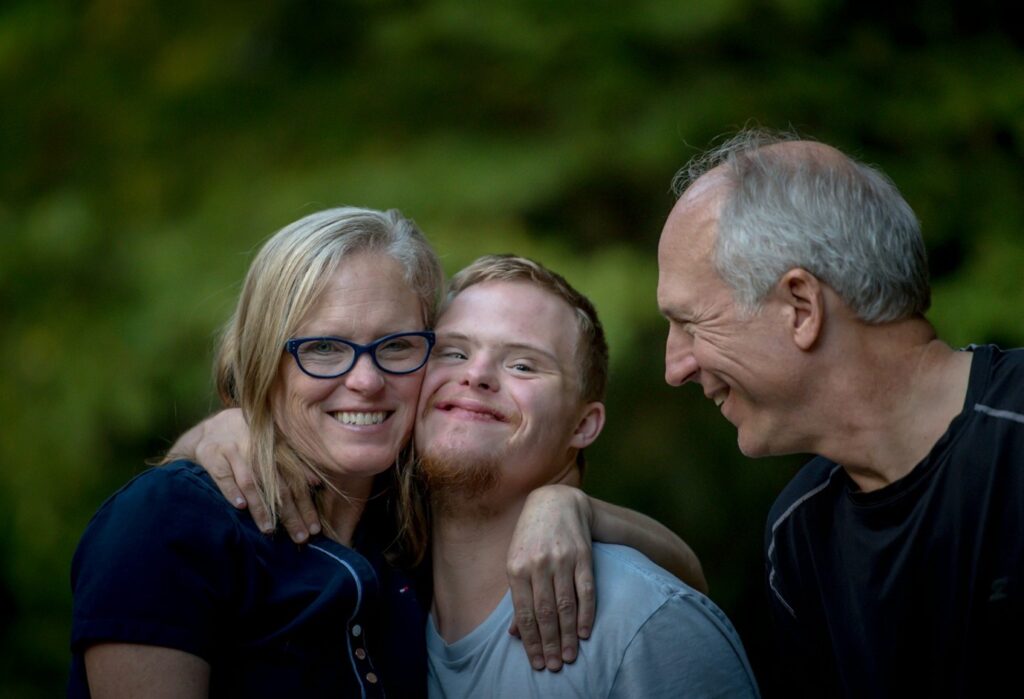 a young man with down-syndrome is stood outside with his parents. he has his face pressed to his mothers. his father is on the other side looking at him. they are all smiling.