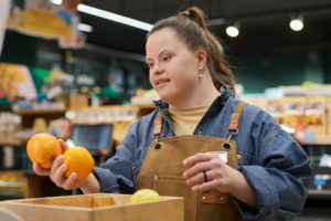 a young woman with down syndrome is working in a supermarket. in her left hand, she is holding two oranges, as if going to put them down in the box in front of her. on her right hand, she has a black payment ring on. she is smiling.