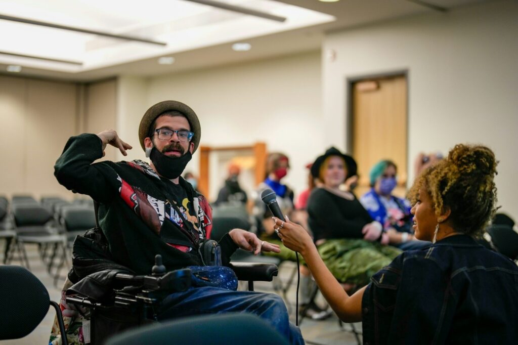 a young man in a wheelchair is looking to the side. a woman with a microphone is sat is knelt in front of him holding the microphone up.