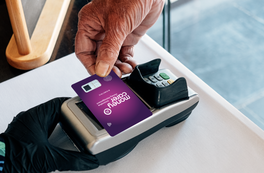 A hand is holding a Money Carer biometric payment card against a payment terminal.