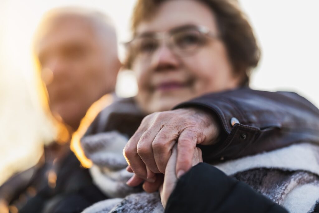 an elderly man and woman are sat outdoors. the woman, in the foreground, is holding someone's hand that is out of shot. the man in the background is blurry.