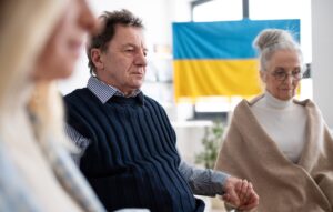 a group of elderly people are sat indoors, holding hands. they are looking sad, and looking down at the floor. in the background, there is a ukranian flag.