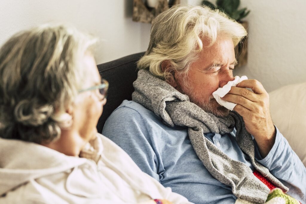 an elderly woman and man are sat indoors. the woman is looking over at the man. the man is blowing his nose into a tissue.