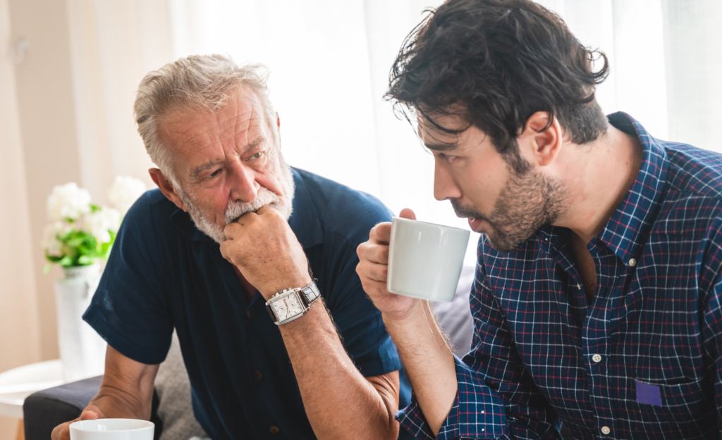 Two men are sat on a sofa drinking out of mugs. They are both looking at each other. The man on the left is older, and looks thoughtful. The man on the right is younger.