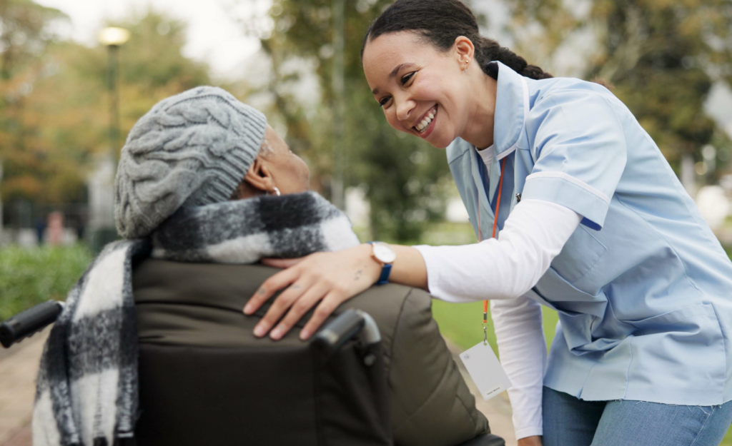 A young woman has her arm on an older woman's shoulder in an outside setting. The older woman is in a wheelchair, facing away from the camera. Both women are looking at each other and smiling.
