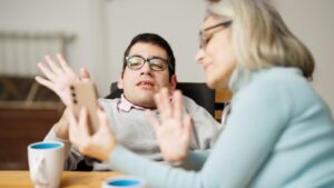Young man with spina bifida holding up hand during a video call on a smartphone with a caregiver. He is wearing glasses. The carer is to his left, and is holding the phone and also waving at the camera.