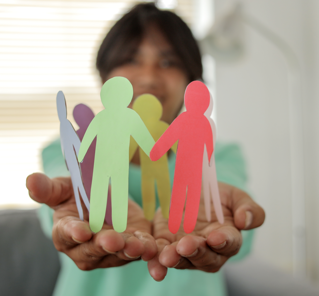 A young woman has her arms outstretched, smiling. Her face is blurry. In her hands, she holds a circle of paper dolls of all different colours.